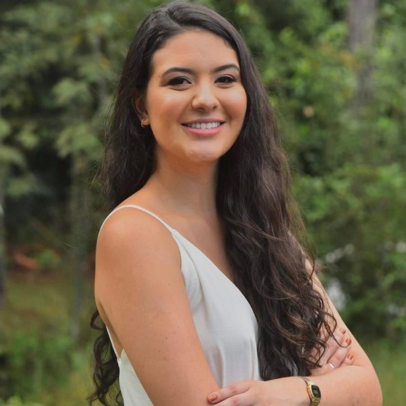 Bianca Caliari sorrindo, de braços cruzados, ao ar livre